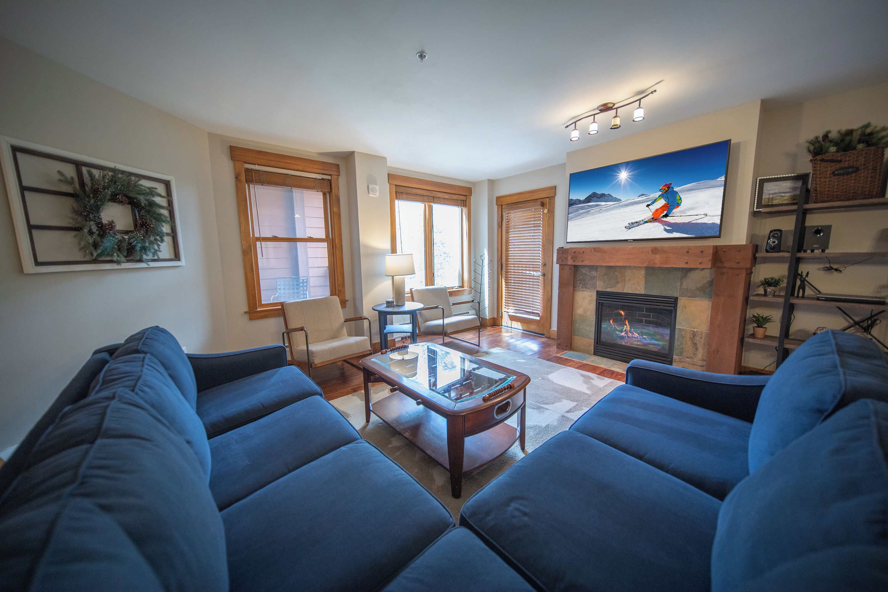 A living room with blue sofas, a glass coffee table, a fireplace, and a painting of a skier.