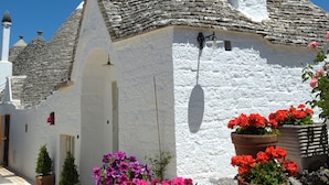 A white building with a stone roof, a chimney, and a small door. There are potted plants with red and pink flowers in front of the building. - Romantic Trulli (Alberobello)