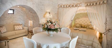A stone-walled room with a round table, white chairs, a sofa, and a window with a view.