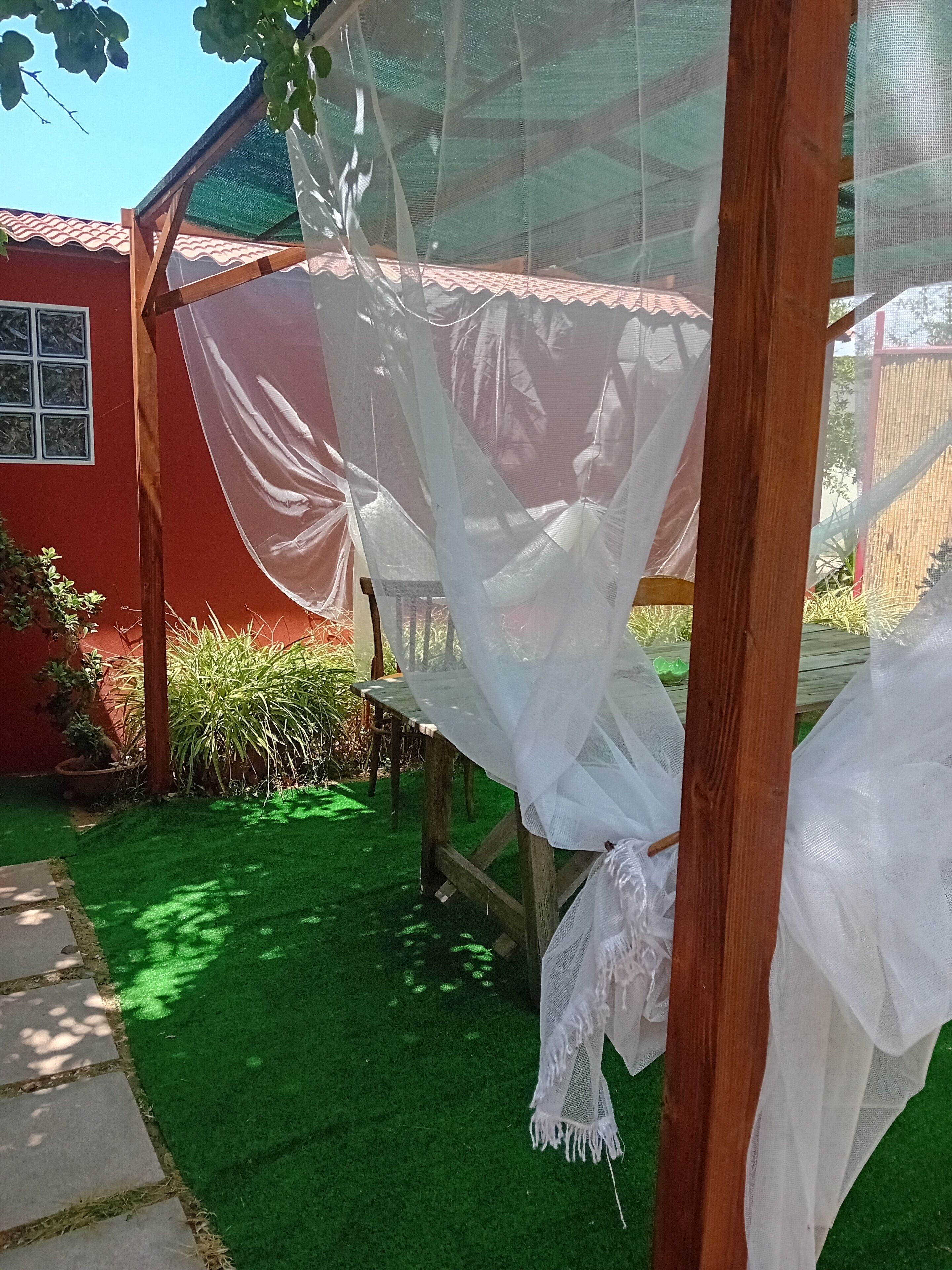 Una estructura de madera con un toldo de malla blanca, rodeada de césped artificial verde y plantas en macetas.