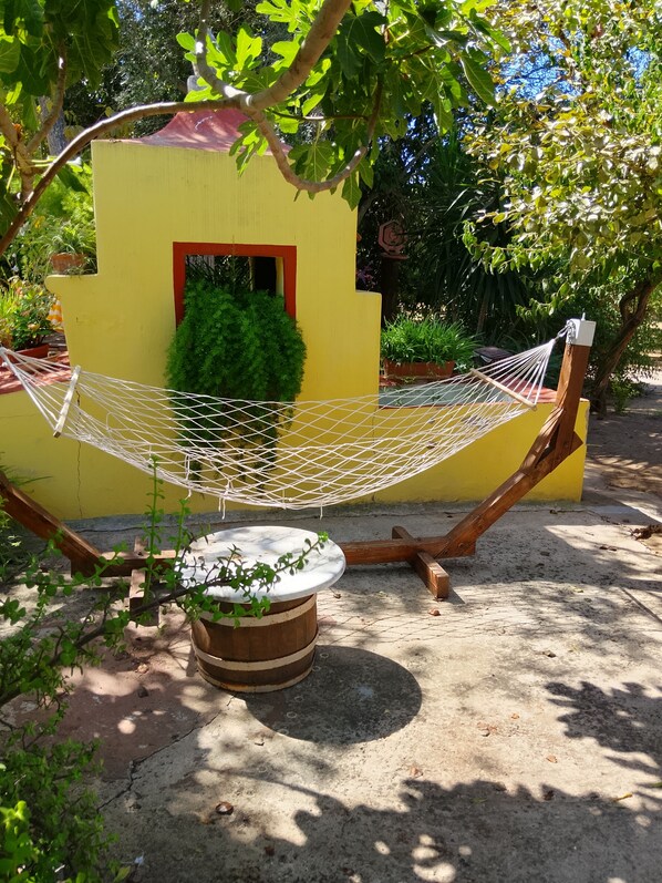 A hammock tied between a yellow wall and a tree, a wooden bench, and a potted plant.