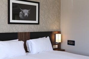 A hotel room with a bed, white pillows, a framed picture of a bull, a night lamp, and an emergency instruction sign.