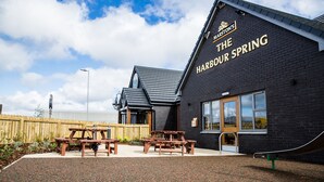 A building named "The Harbour Spring" with a black exterior, wooden picnic tables, and a wooden fence in the background. - Harbour Spring, Peterhead by Marston's Inns (Peterhead)