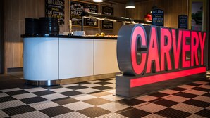 A restaurant counter with a large sign reading "CARVERY" and menu boards advertising dishes like "GOLARGE" and "TRULY ROASTED PIGS".