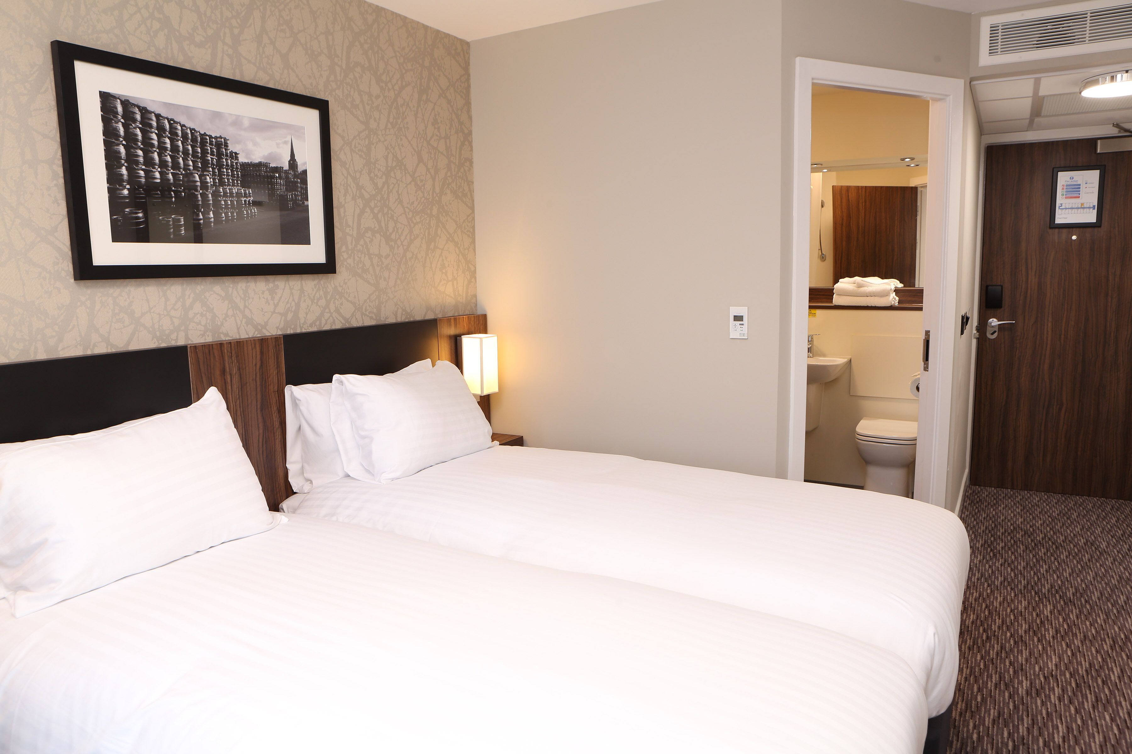 A hotel room with a large bed, a framed picture on the wall, a door leading to a bathroom, and a carpeted floor.