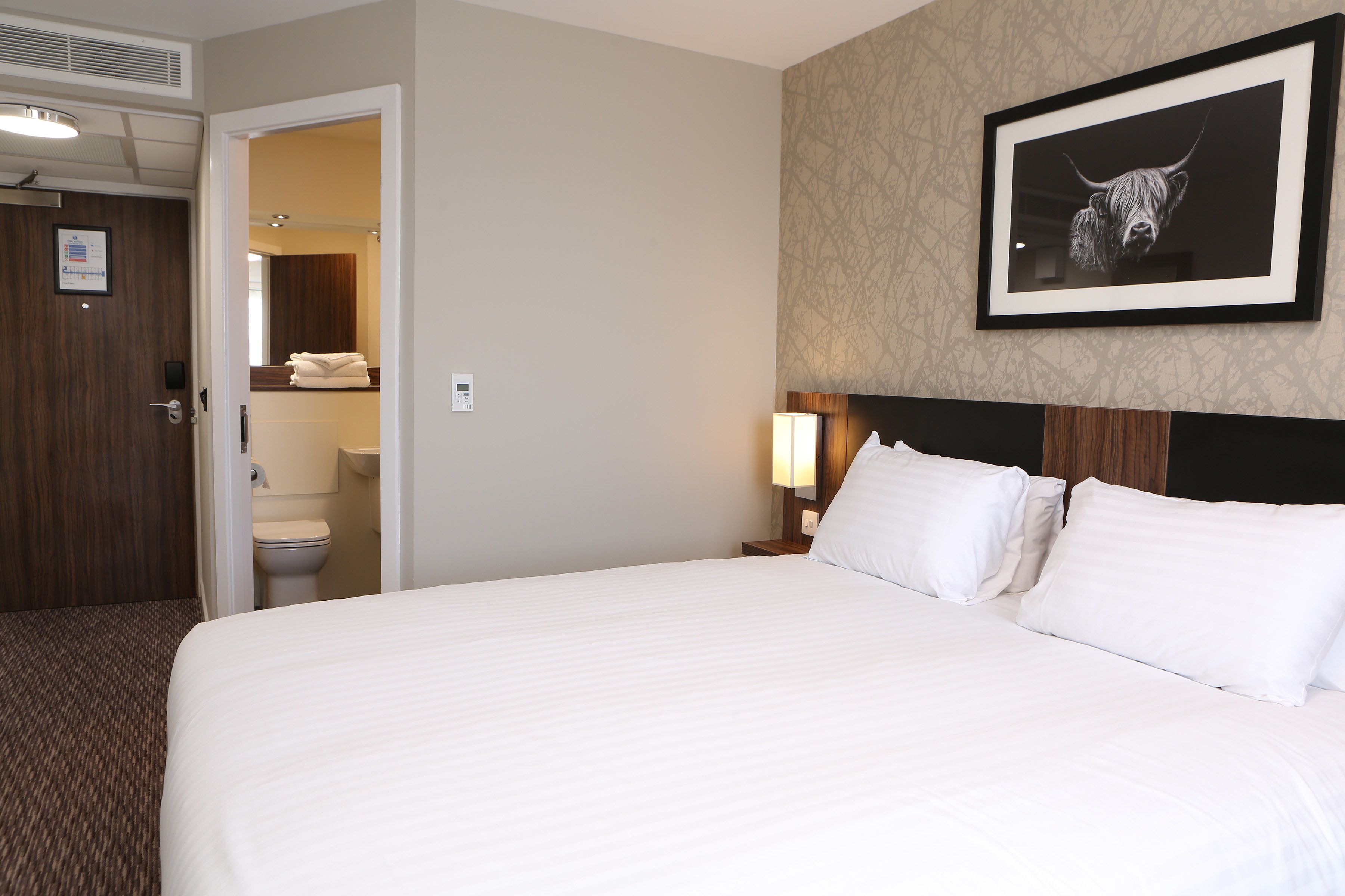 A hotel room with a large bed, a headboard with a framed picture, a bedside lamp, and a view of the bathroom.