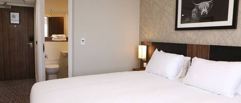 A hotel room with a large bed, a headboard with a framed picture, a bedside lamp, and a view of the bathroom.