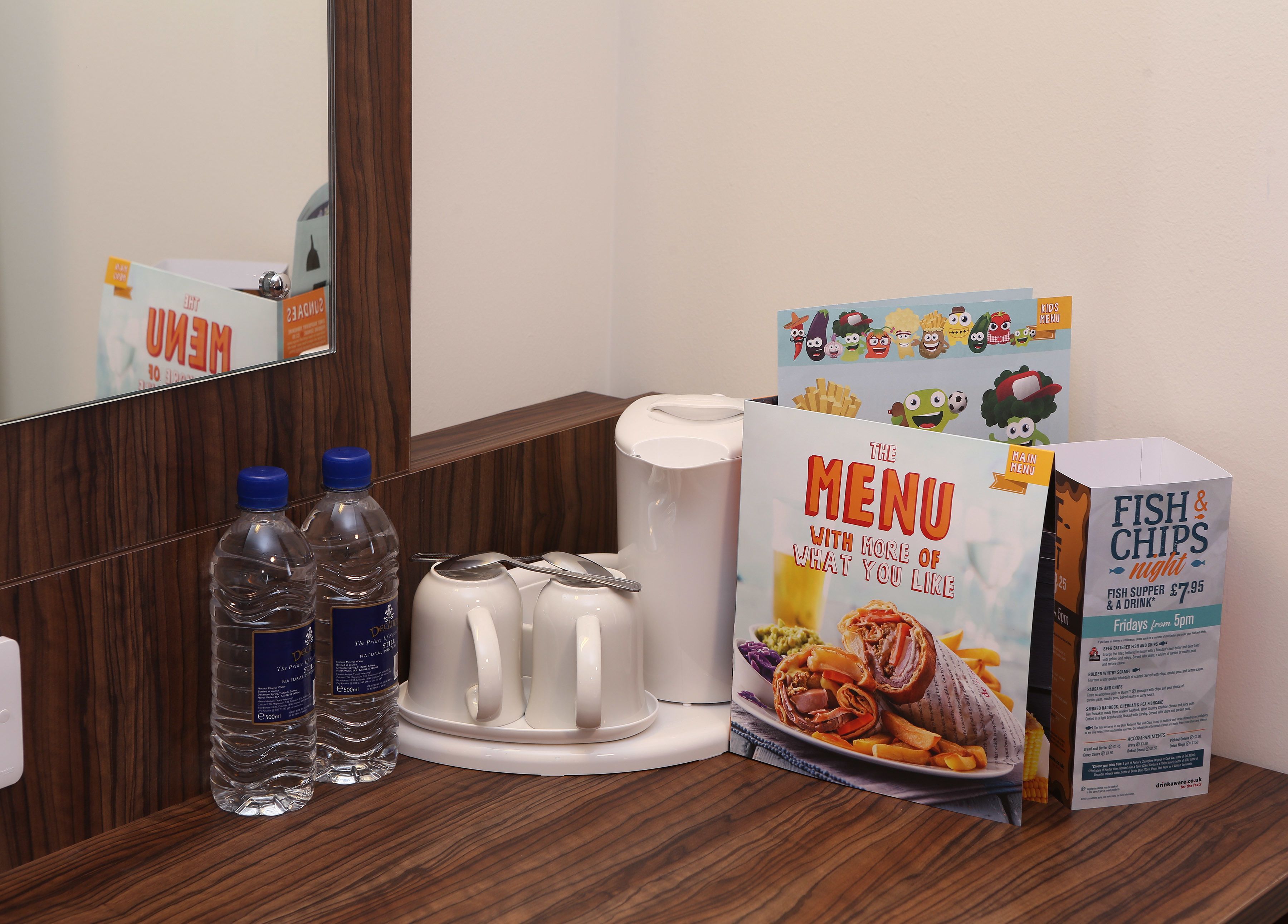 A hotel room with a wooden floor, a mirror, a white coffee maker, two water bottles, a menu stand, and a box of fish & chips.