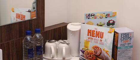 A hotel room with a wooden floor, a mirror, a white coffee maker, two water bottles, a menu stand, and a box of fish & chips.