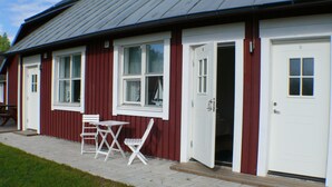 Un bâtiment rouge avec des fenêtres et des portes à encadrement blanc, un petit espace extérieur aménagé avec des chaises et une table, tous deux de couleur blanche, et un chemin pavé.