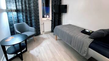 Een kleine, moderne kamer met een bed, een stoel, een tafeltje, een televisie en een raam met gordijnen.