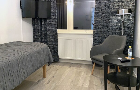 A room with a bed, a chair, a small table, a TV mounted on the wall, and a window with curtains. - Vara Pensionat (Vara)