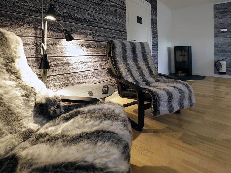 A cozy room with a wooden wall, a black chair with a fur throw, a small table with a lamp, and a fireplace.