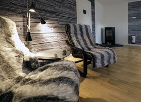 Um quarto acolhedor com parede de madeira, uma cadeira preta com manta de pele, uma mesinha com abajur e uma lareira.