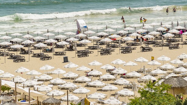 Ein Strand mit vielen Sonnenschirmen und Liegestühlen, ein sandiger Uferstreifen und das Meer im Hintergrund.