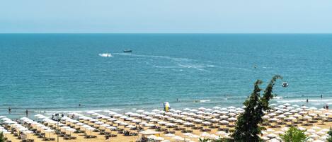 A beach with rows of umbrellas and sun loungers, a clear blue sea, and a few boats in the distance.