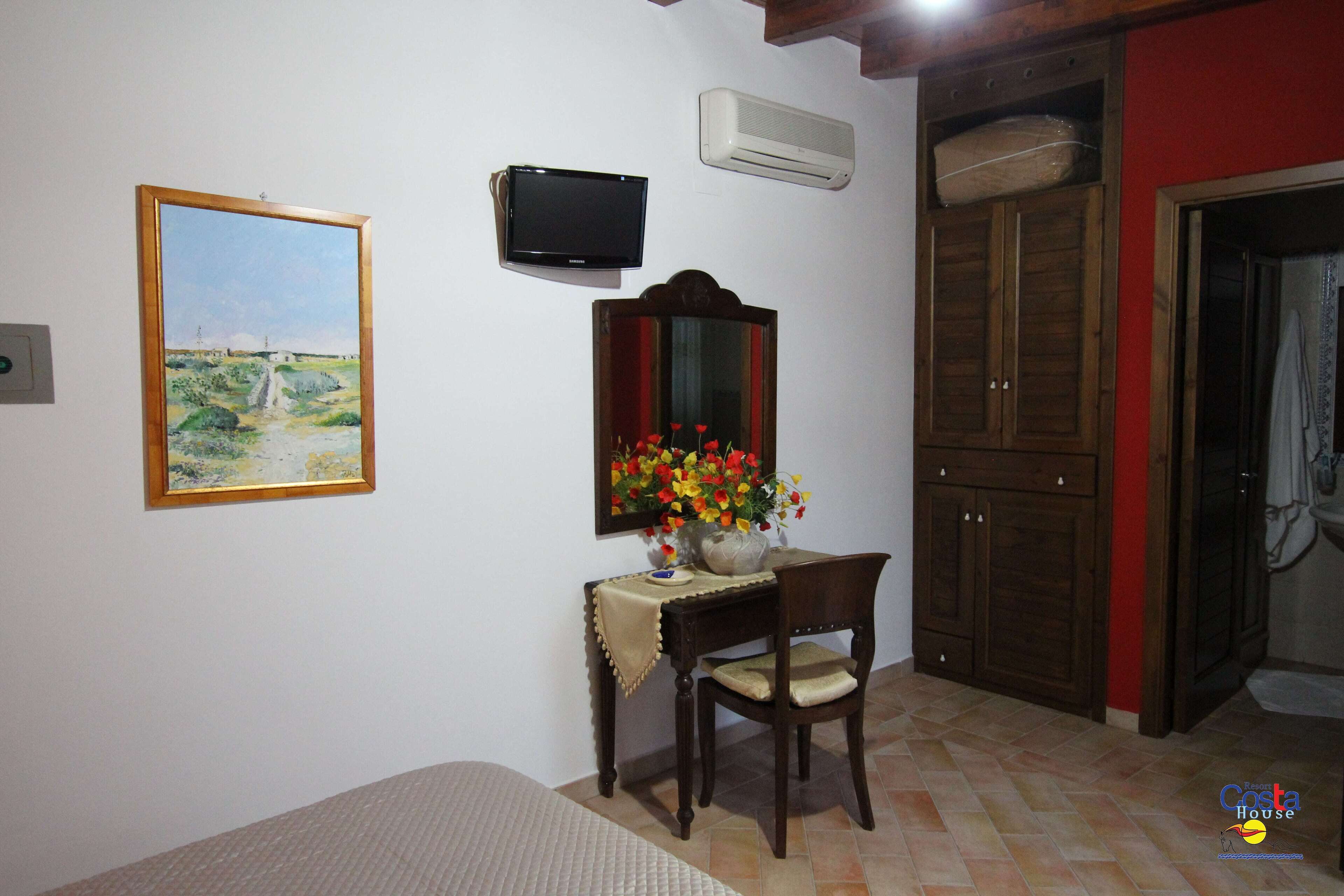 A room with a bed, a desk with a chair, a painting, a television, an air conditioner, and a wardrobe.