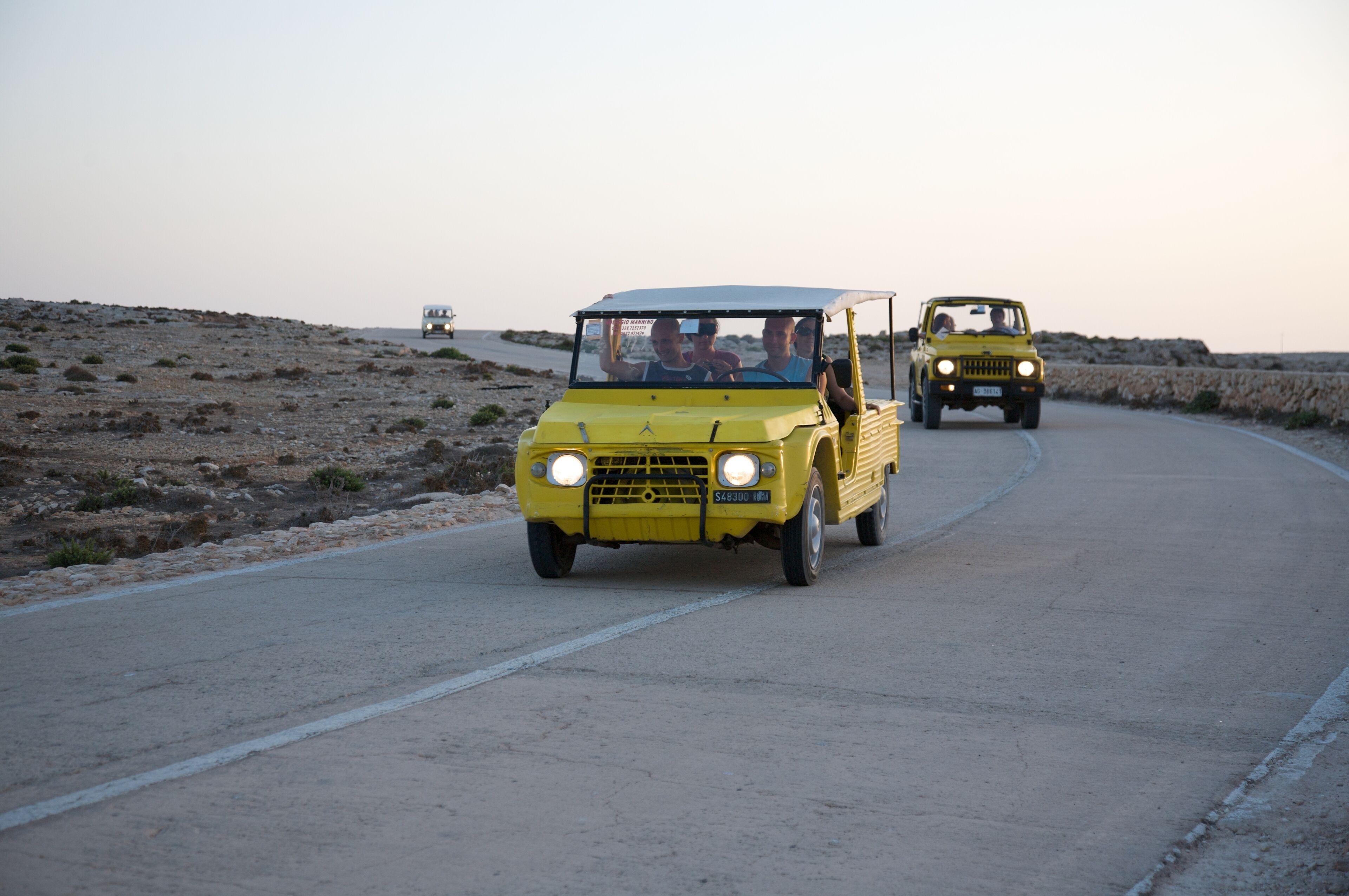 Two yellow vehicles on a paved road with a stone wall on one side and a rocky landscape on the other.