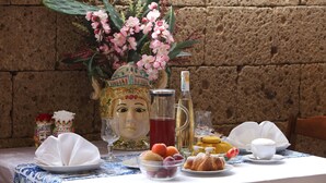 Mesa posta com refeição, incluindo frutas, doces e uma bebida, com parede de pedra ao fundo.