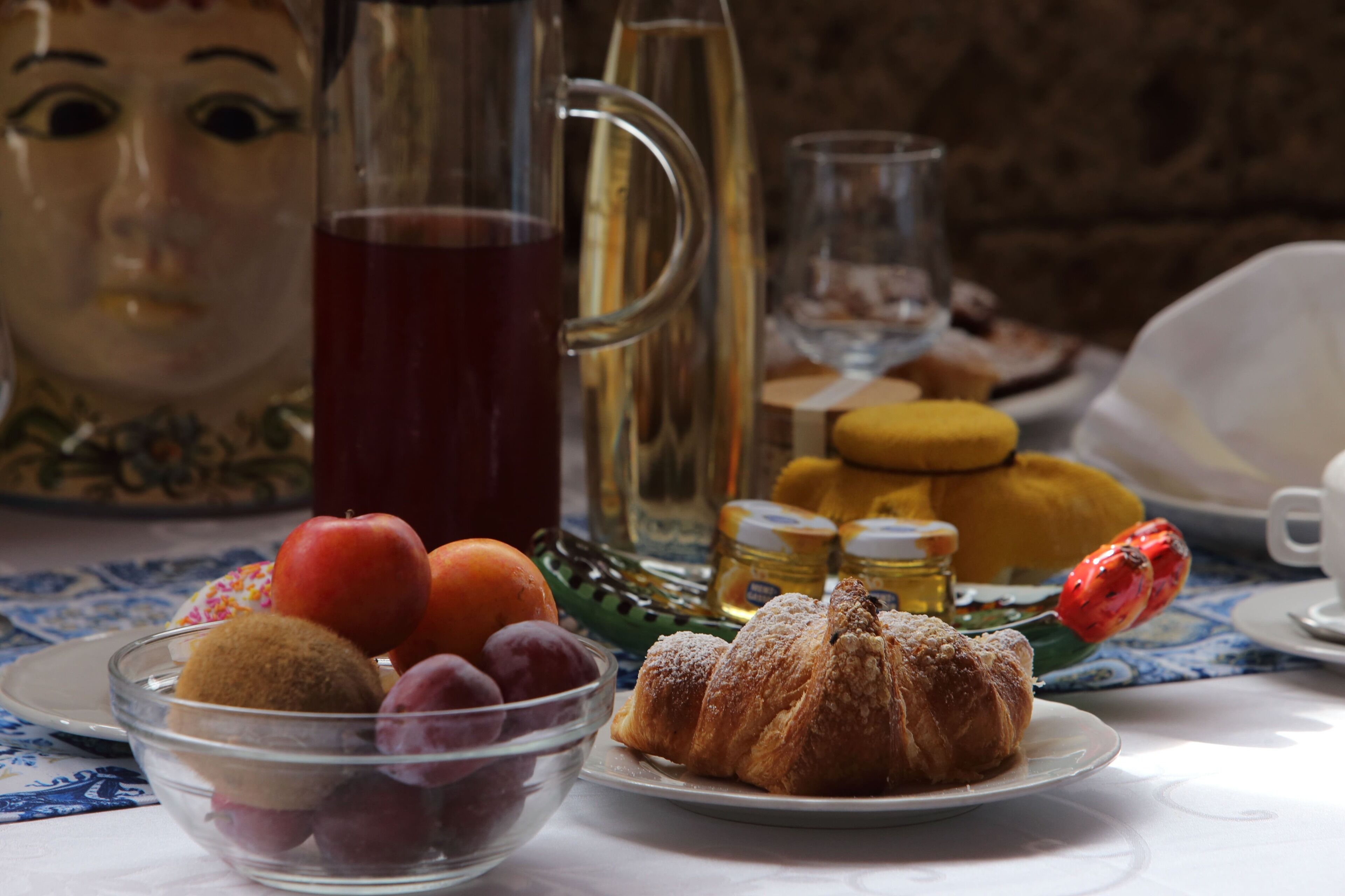 Una mesa con un bol de vidrio lleno de fruta, un plato con un pastel y una jarrón de líquido rojo.