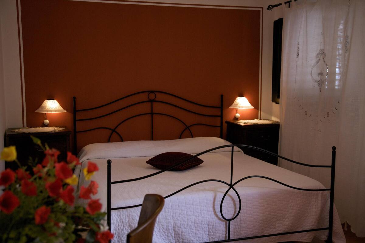 Un letto con struttura in metallo, due lampade sui comodini e una finestra con tende leggere.