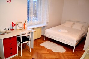 Quarto com cama, escrivaninha com cadeira, radiador e janela com vista para edifícios.