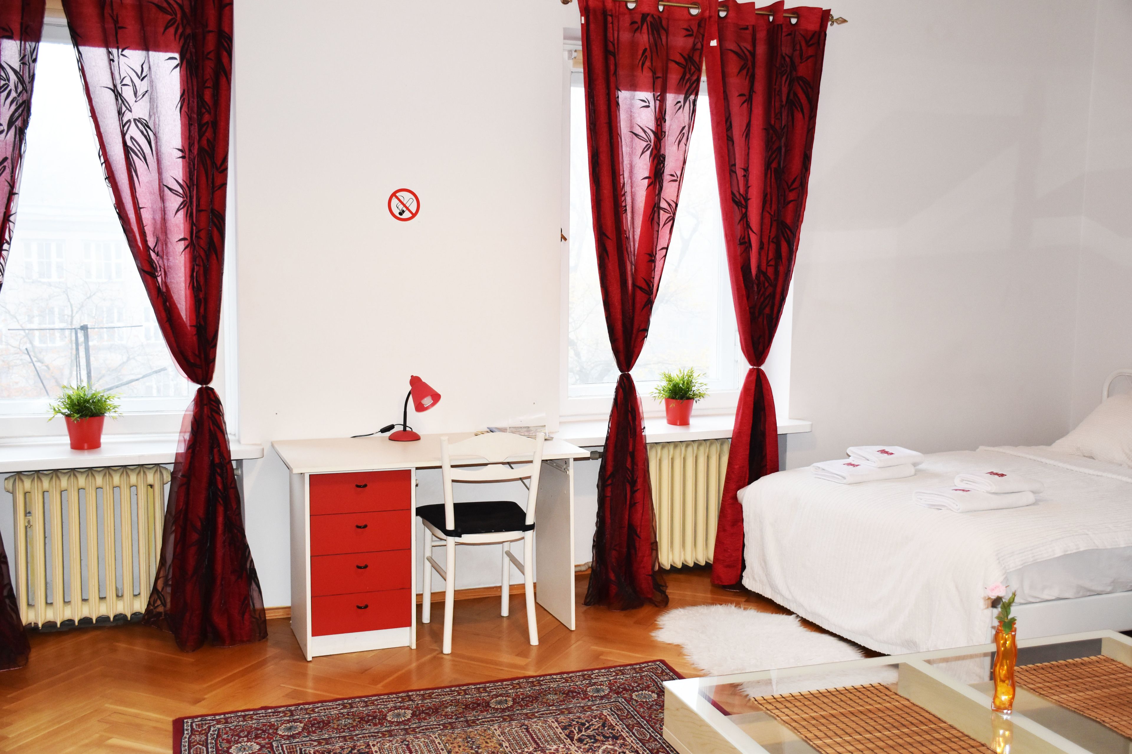 Une chambre à coucher avec un lit blanc, un bureau avec une chaise, un radiateur et deux plantes en pot.