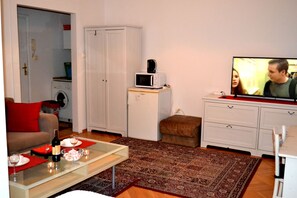 En kompakt opholdsstue med sofa, sofabord med flaske og glas, mikroovn, fjernsyn og vaskemaskine i baggrunden.