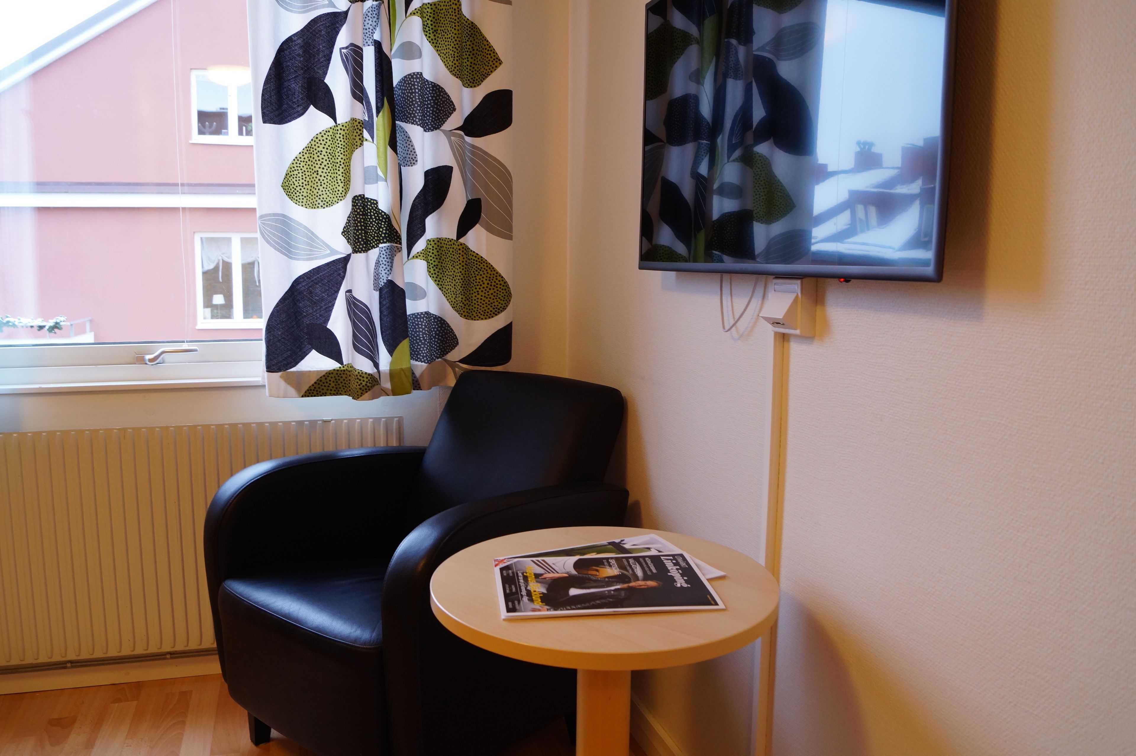 Ett rum med en svart läderfåtölj, ett litet runt bord med en tidning, en tv som är monterad på väggen och ett fönster med en gardin.
