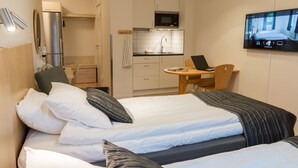 Une chambre d’hôtel avec deux lits, un bureau avec un ordinateur portable, un téléviseur fixé au mur et une kitchenette.