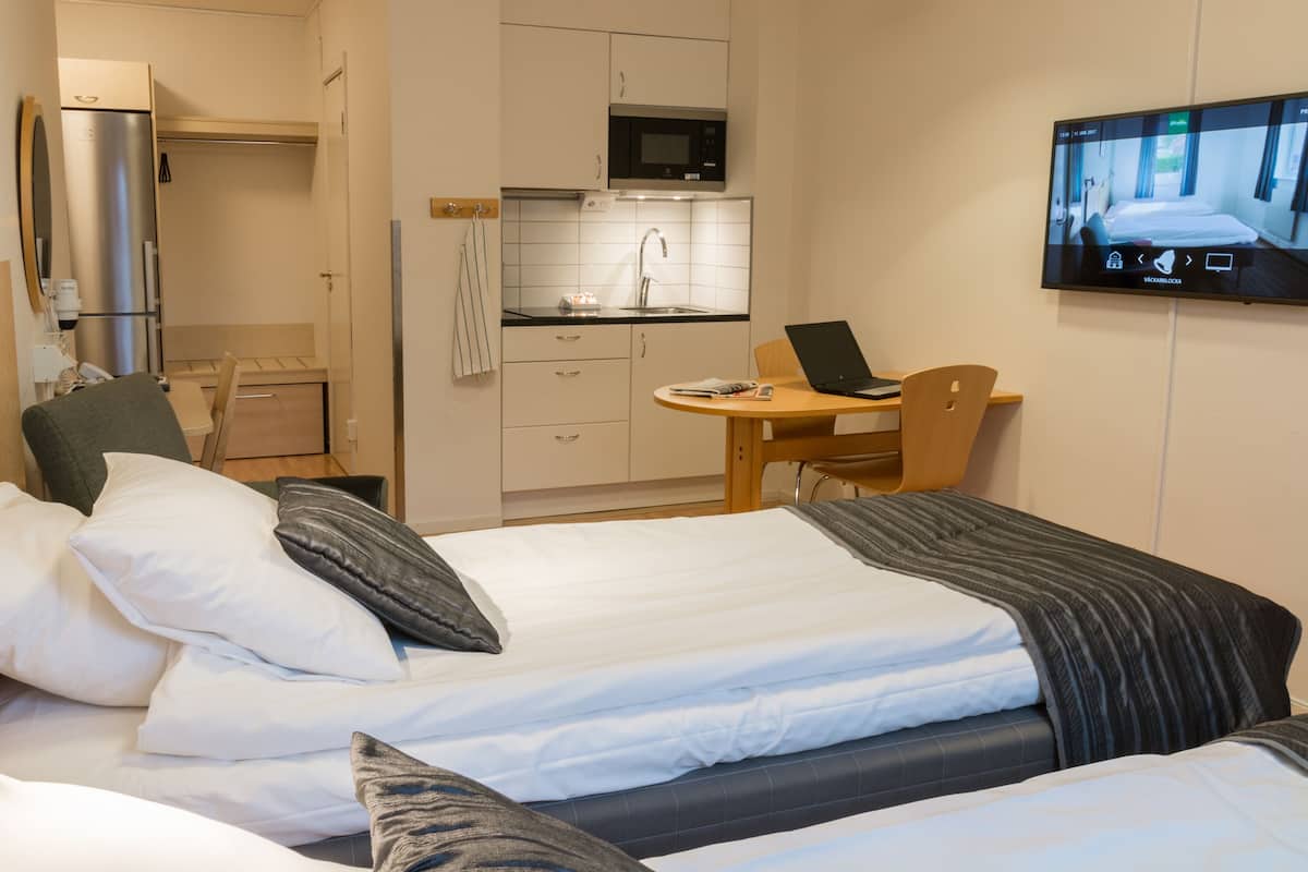 Habitación de hotel con dos camas, un escritorio con una computadora portátil, un televisor en la pared y una kitchenette.