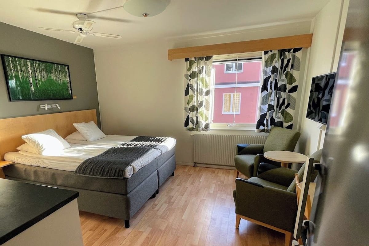Un dormitorio con una cama, una silla, una mesa, un ventilador de techo y una ventana con cortinas.