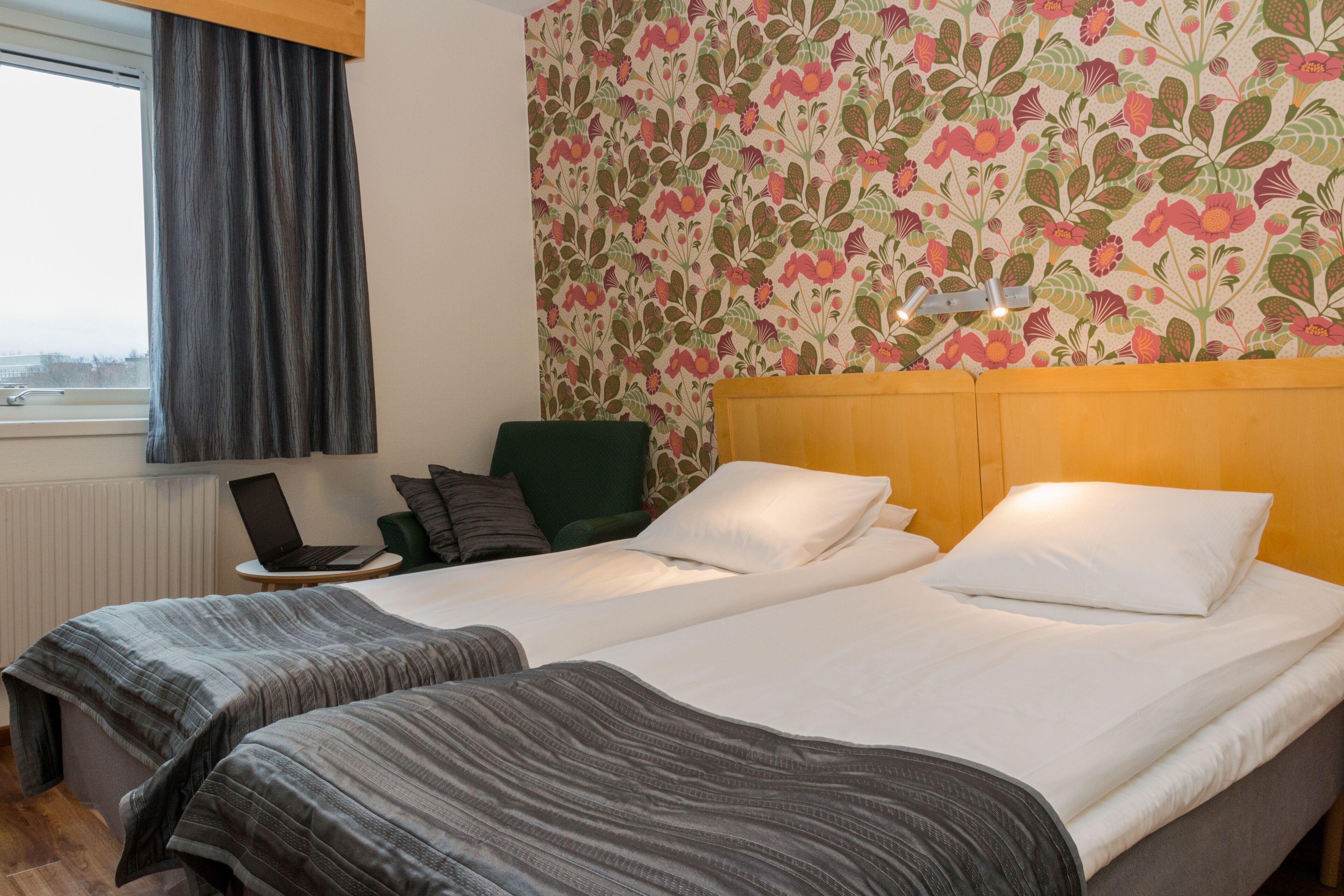A hotel room with two beds, a laptop on a small table, a green chair, and a window with curtains.
