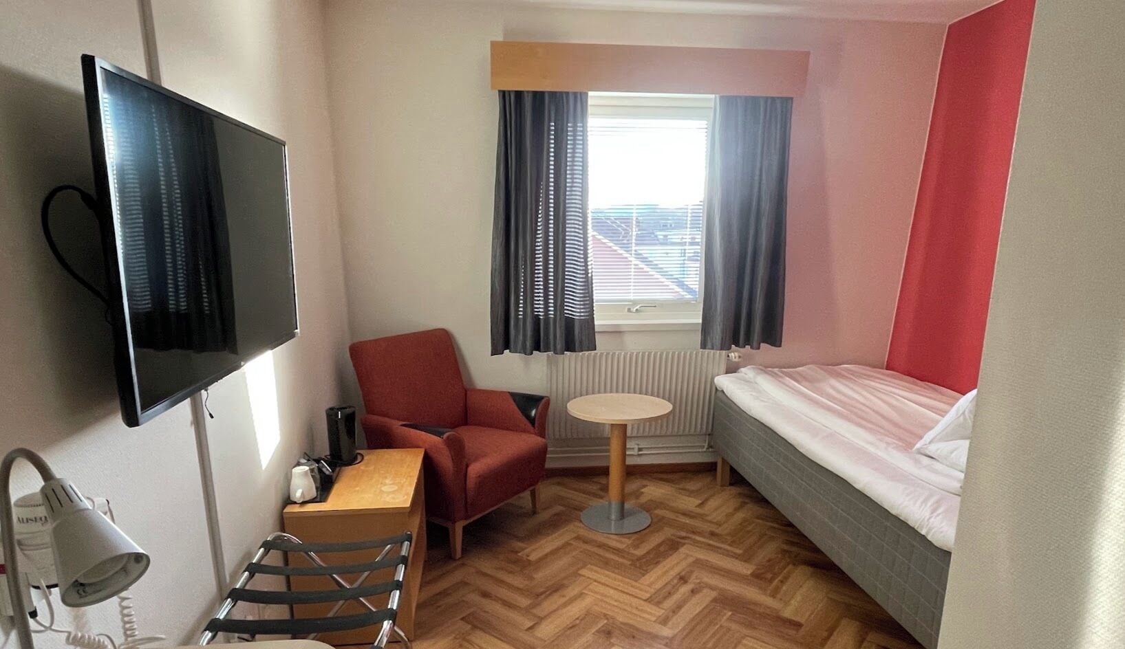 A compact hotel room with a bed, a desk with a chair, a television, and a radiator.