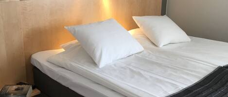 Un lit bien fait, avec du linge de lit blanc et des oreillers, une table de chevet sur laquelle se trouvent un livre et une télécommande, et un luminaire fixé au mur.