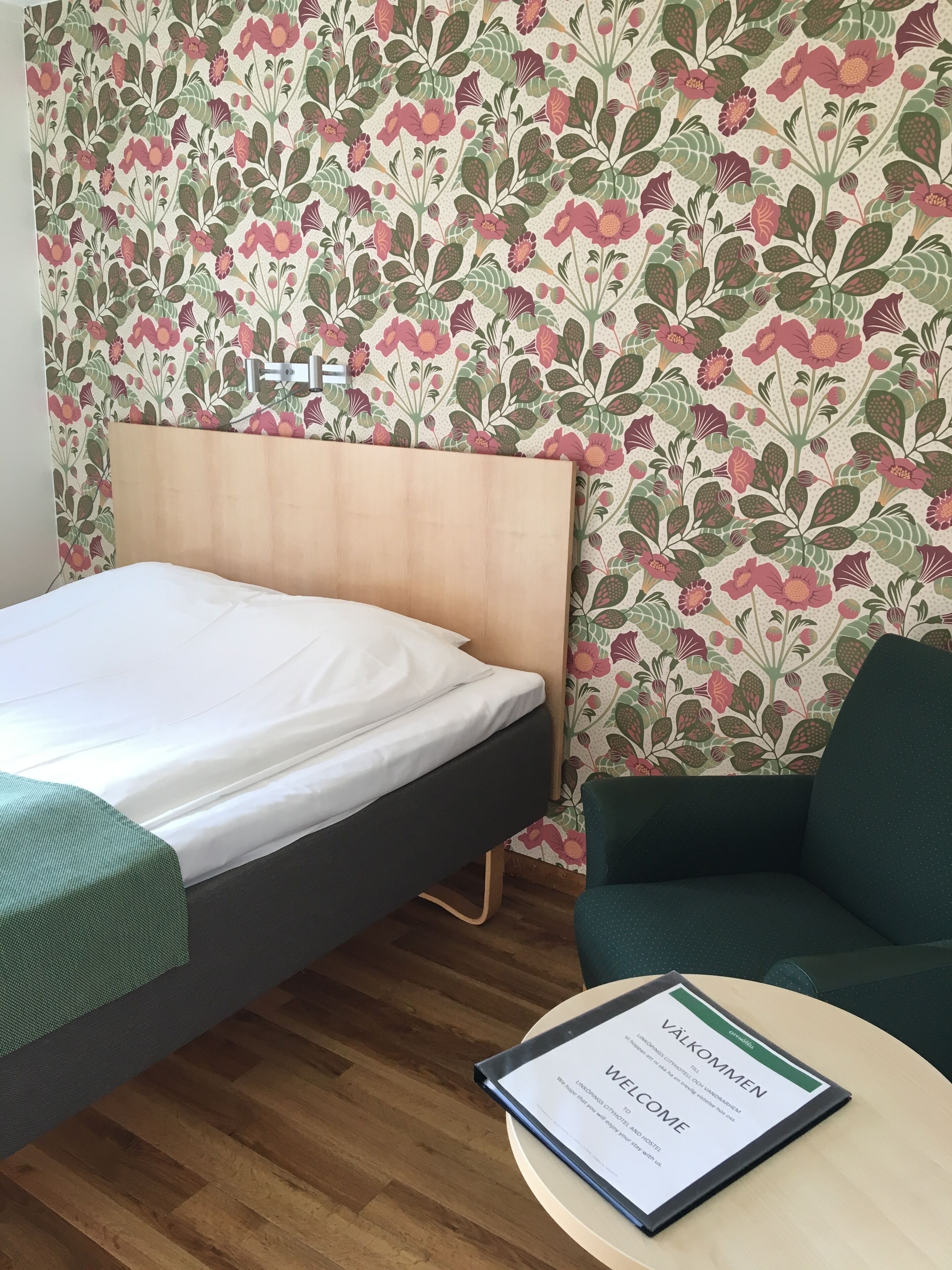 Una habitación de hotel con una cama, un sillón verde y un letrero de bienvenida.