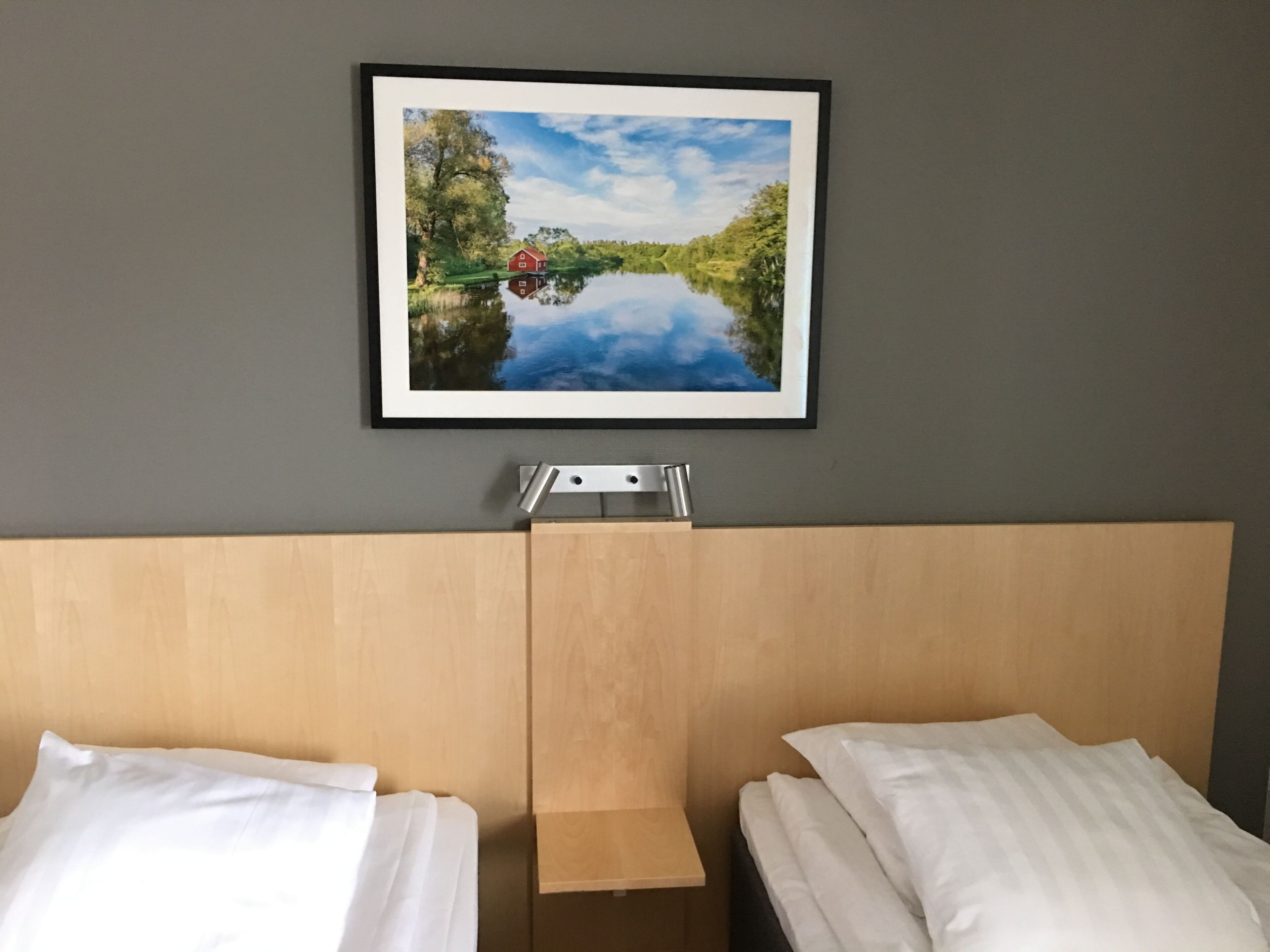 Habitación de hotel con una cama, una pequeña mesa de madera y un cuadro enmarcado con una imagen de un lago y una casa.