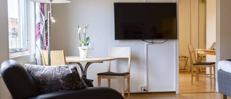 A modern hotel room with a sofa, a small table with a vase of flowers, a flat-screen TV mounted on the wall, and a desk with a chair.