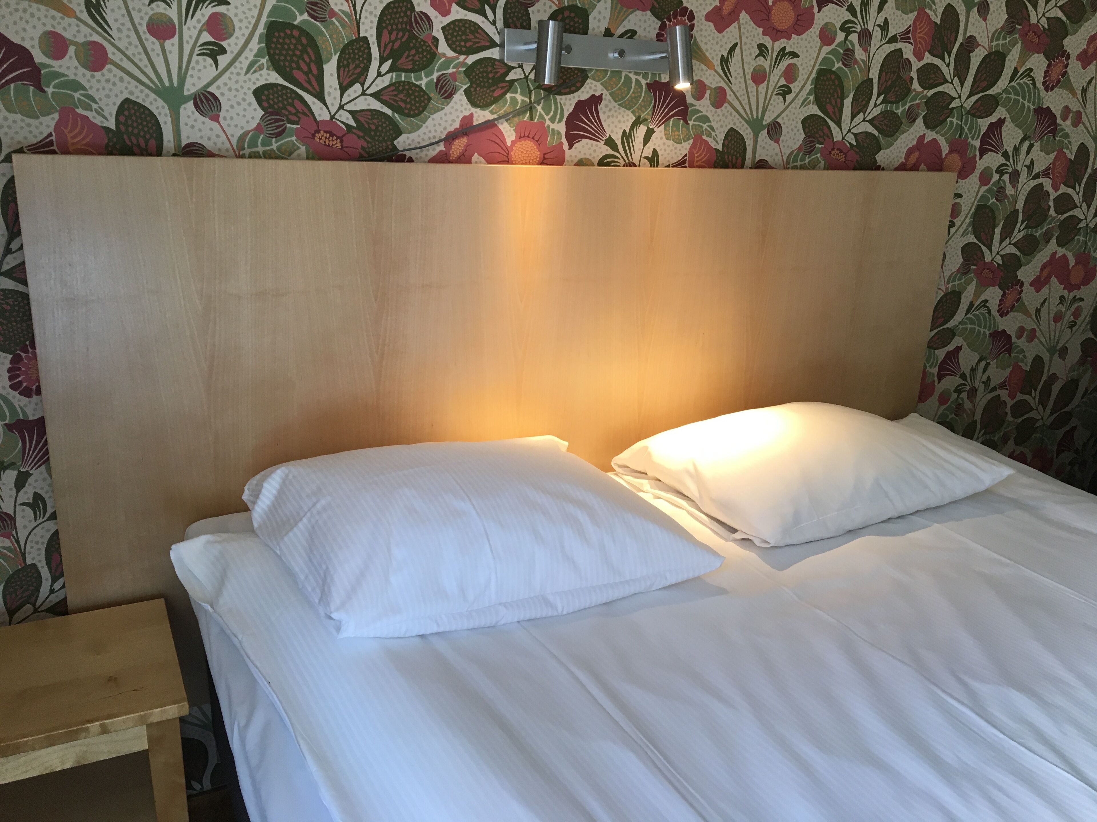 Una cama bien hecha con sábanas y almohadas blancas, un cabecero de madera y una mesita de noche con una lámpara.