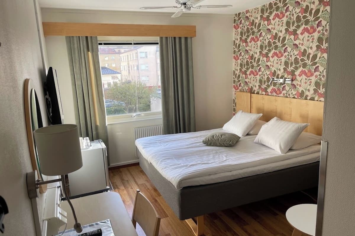 Una habitación de hotel con cama, escritorio con lámpara, silla, una mesita y ventana con cortinas.