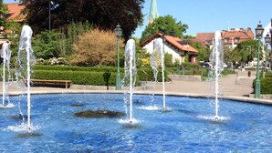 Une fontaine dotée de plusieurs jets d’eau, située sur une place publique et entourée de verdure ainsi que de bâtiments à l’architecture traditionnelle.