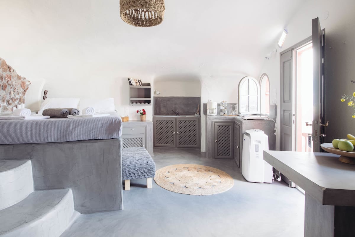 A modern bedroom with a large bed, a bedside table, and a small seating area. There is a kitchen area with a sink, stove, and cabinets.