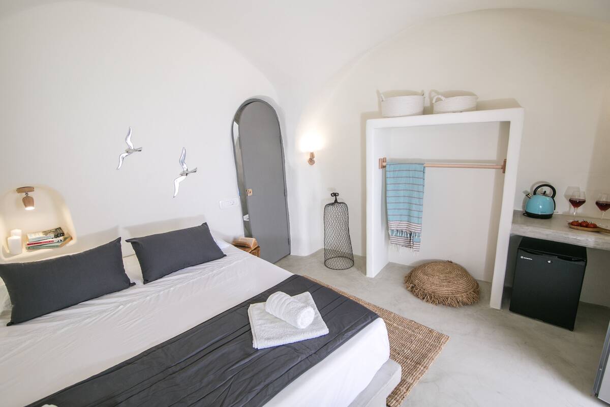 A bedroom with a bed, pillows, a bedside table with a lamp, a small stool, a basket, and a towel on the bed.
