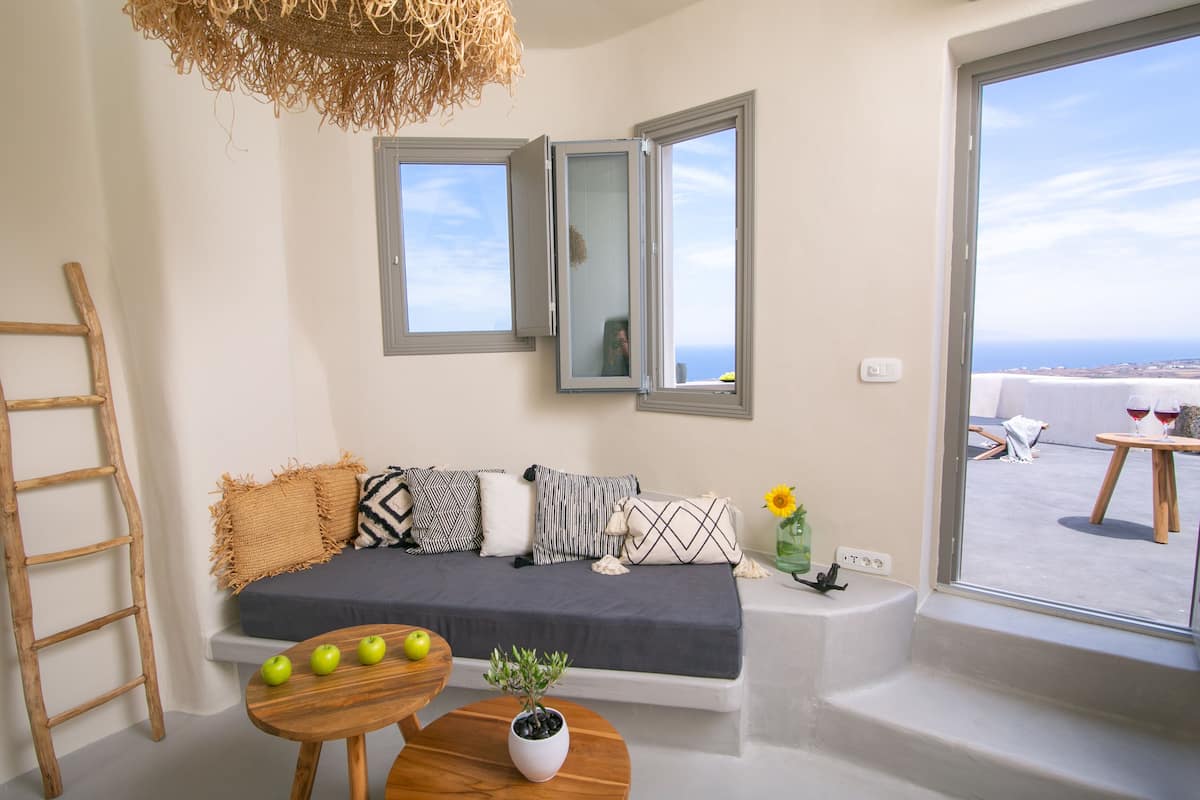 A modern living room with a sofa, a wooden table with apples, a potted plant, and a view of the sea through large windows.