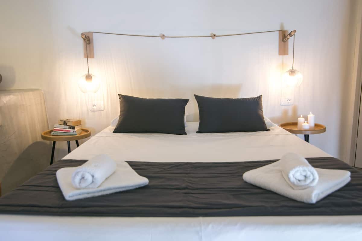 A neatly made bed with two pillows, a bedside table with a book and a candle, and a wall-mounted light fixture.