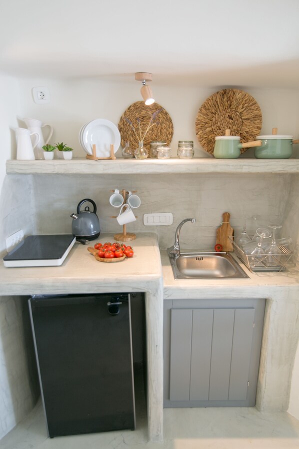 Una cocina compacta con lavavajillas integrado, un fregadero y una encimera con un hervidor, una tabla de cortar con tomates y un refrigerador pequeño.