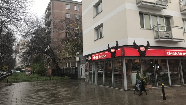 A steakhouse with a red and white sign, located at the corner of a street with a brick sidewalk. - West Apartments Hoża 36 (Warsaw)