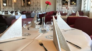 A restaurant with tables set for dining, featuring clear glassware, a vase with a red flower, and a patterned wallpaper interior.