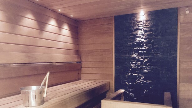A sauna with wooden benches, a stone wall, and a metal bucket.
