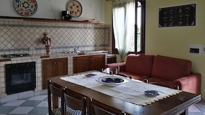 A dining area with a table set for two, a kitchen with tiled backsplash, and a red sofa in the living area. - Villa La Rocca (Castelsardo)